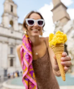 K. HEBDA olejek cytrynowy Kobieta w białych okularach trzyma żółte lody na tle miejskiej architektury