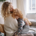 Szczęśliwa mama w jasnym swetrze czule obejmuje i całuje w głowę swoje rudowłose dziecko na przytulnej kanapie, budując atmosferę spokoju i bezpieczeństwa.