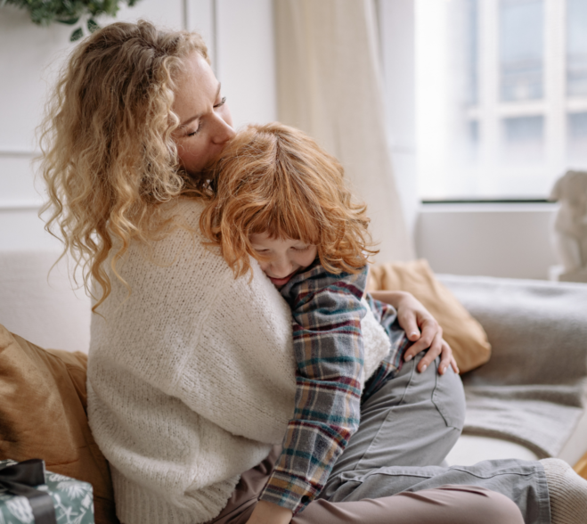 Szczęśliwa mama w jasnym swetrze czule obejmuje i całuje w głowę swoje rudowłose dziecko na przytulnej kanapie, budując atmosferę spokoju i bezpieczeństwa.