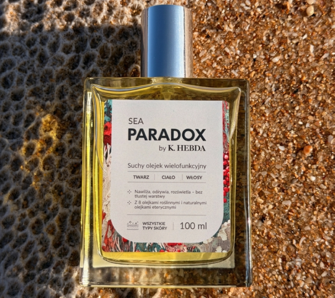 Flakon luksusowego suchego olejku wielofunkcyjnego Sea Paradox by K. Hebda o pojemności 100 ml, ustawiony na naturalnym piasku plaży i obsypany kryształami soli morskiej.