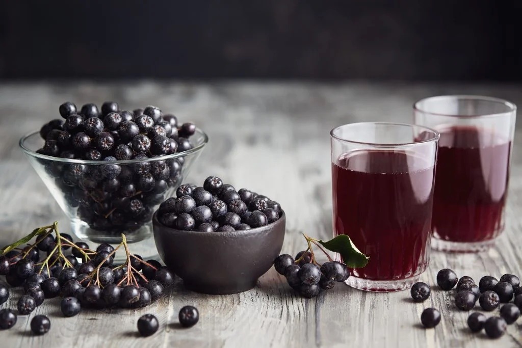 aronia na odporność Miseczki z owocami aronii i dwie szklanki soki o ciemnoczerwonym kolorze na drewnianym blacie w otoczeniu małych czarnych owoców