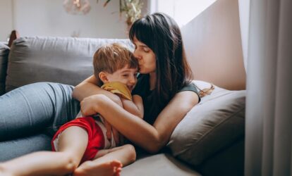Mama i syn przytulają się na szarej kanapie, mama całuje syna w czoło