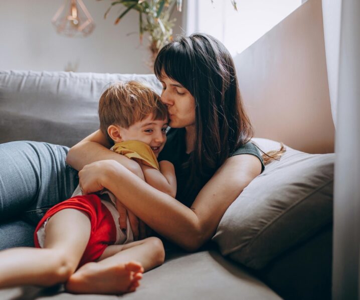 Mama i syn przytulają się na szarej kanapie, mama całuje syna w czoło