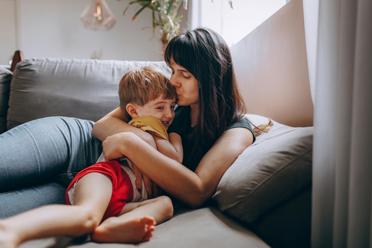Olejki eteryczne na katar u dzieci Mama i syn przytulają się na szarej kanapie, mama całuje syna w czoło