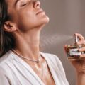 Elegancka kobieta w brązowym swetrze rozpylająca zapach z flakonu perfum z serii Paradox na punkty tętna na szyi.