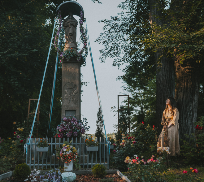Tradycyjna polska kapliczka przydrożna otoczona kwiatami i potężnymi drzewami, obok których stoi autorka zapachu woda toaletowa Lipa z miodem.