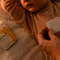 Zbliżenie na dłoń rodzica naklejającą aromaterapeutyczny Plaster Lekki Oddech K. Hebda na ubranko śpiącego dziecka obok kartonowego opakowania z żółtym słonikiem.