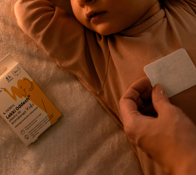 Zbliżenie na dłoń rodzica naklejającą aromaterapeutyczny Plaster Lekki Oddech K. Hebda na ubranko śpiącego dziecka obok kartonowego opakowania z żółtym słonikiem.